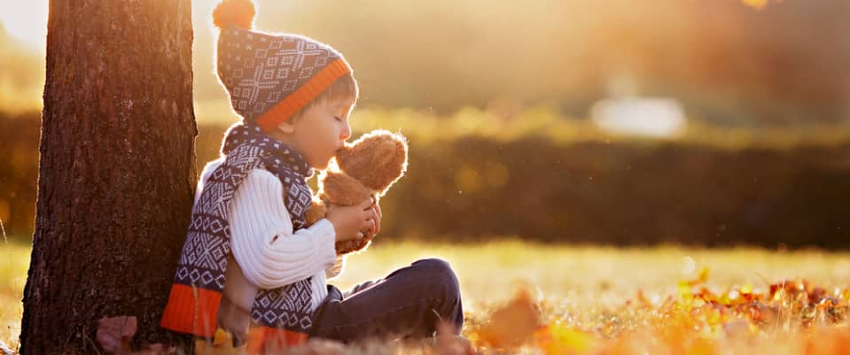 little boy sitting under a tree while kissing his bear stuffed toy 